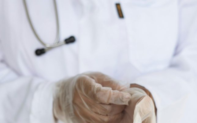 Faceless ethnic medical worker in lab coat and stethoscope taking of transparent gloves after approaching patients for examination in modern hospital